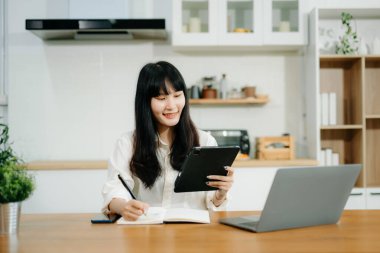 Evde otururken dizüstü bilgisayar kullanan bir kadın. Genç bir kadın mutfakta oturuyor ve dizüstü bilgisayarla çalışıyor.