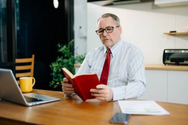 Olgun iş adamı evde otururken dizüstü bilgisayar kullanıyor. Adam mutfakta oturuyor ve dizüstü bilgisayarla çalışıyor.