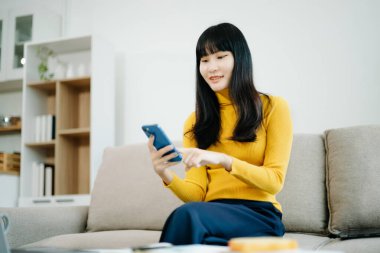 Genç Asyalı kadın rahat bir kanepede rahat rahat oturup akıllı telefondan mesajlaşıyor, cep telefonu kullanıyor, internette mesajlaşıyor, evdeki kanepede online alışveriş yapıyor.