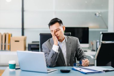 Ofis masasında dizüstü bilgisayarla oturan yorgun işadamı, baskı ve şirket baskısı altında yoğun ve stresli hissediyor..