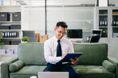 Modern bir ofiste tabletle çalışan Asyalı işadamı belgelere bakarken güvenle gülümsüyor..