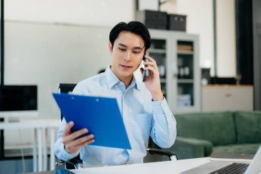 Asyalı İşadamı telefonda konuşuyor ve ofiste otururken gülümseyen bir laptop kullanıyor.