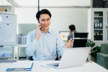 Asyalı İşadamı telefonda konuşuyor ve ofiste otururken gülümseyen bir laptop kullanıyor.
