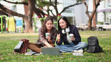 İki kız öğrenci bir dizüstü bilgisayarla çalışırken parkta çimlerin üzerinde oturuyorlar.