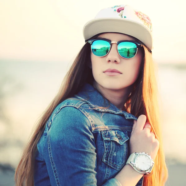 Retrato de moda ao ar livre de menina elegante swag, vestindo boné swag, óculos de sol na moda e jaqueta de ganga, vista incrível da praia ao pôr do sol . — Fotografia de Stock