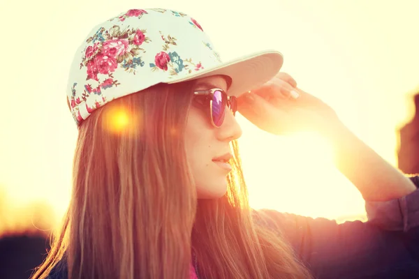 Utomhus mode porträtt av eleganta swag flicka, klädd i swag cap, trendiga solglasögon och jeansjacka, fantastisk utsikt över stranden i solnedgången. — Stockfoto