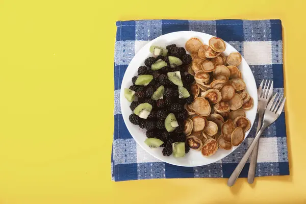 在盘子里放有黑莓和猕猴桃片的迷你煎饼 在黄色背景的蓝色亚麻布餐巾纸上放有两个叉子 — 图库照片