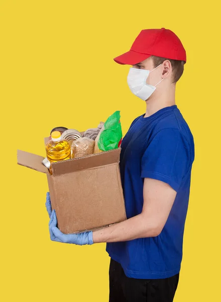 赤い帽子の若い男性ボランティアは黄色の背景に食料品の箱を持っています 社会的概念 貧しい人々のために助けてください 商品の配送 — ストック写真