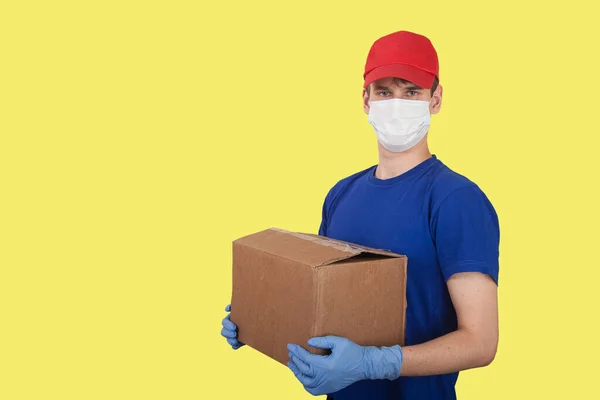 A male volunteer in a medical mask and rubber gloves holds a box on a yellow background. Social help. Delivery of goods by courier. Copy spaes