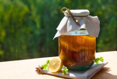 Cam bir kavanozda soğuk bir Kombucha yeşil bir bahçenin arka planına karşı tahta bir masanın üzerinde duruyor. Kürekleri kopyala
