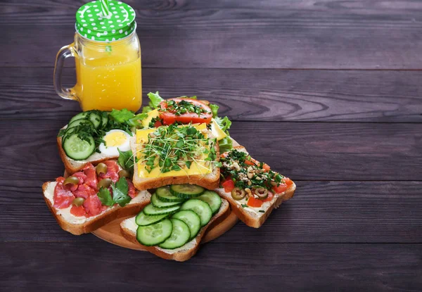 Somonlu, lahanalı, domatesli, salatalıklı, otlu, fındıklı, zeytinli ve taze portakal suyu olan çeşitli lezzetli sandviçler kahverengi ahşap arka planda. Sağlıklı gıda konsepti. Boşluğu kopyala