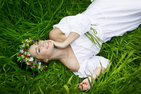 Fille souriante couchée sur l'herbe dans une robe blanche avec une couronne sur la tête — Photo