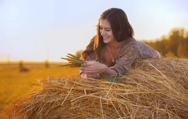 Sonriendo hermosa chica en el heno viendo la hierba de trigo al atardecer — Foto de Stock