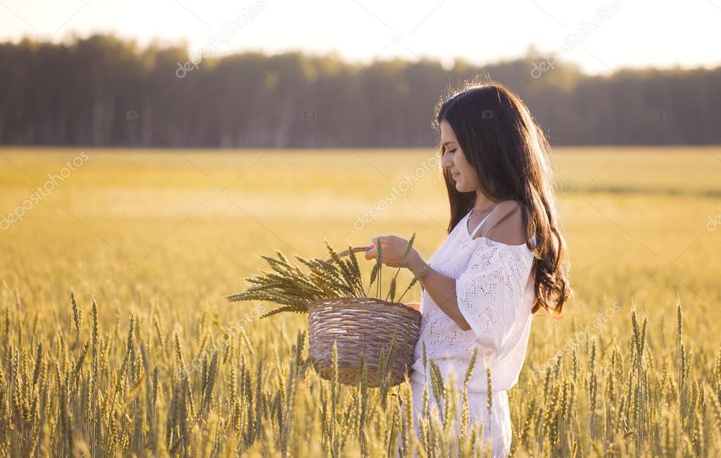 Beautiful brunette in a wheat field looks at the basket with ears