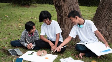 Üç çocuk yeşil çimen zemininde oturuyor, tuvale renk katıyor ve konuşuyor, mutlu bir şekilde birlikte hareket ediyorlar, parkta.