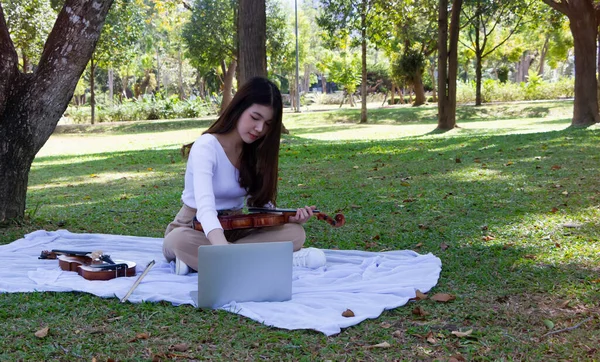 データを検索するためにラップトップを使用して手にバイオリンを持っている若い美しい女性 オンライン学習 公園で — ストック写真