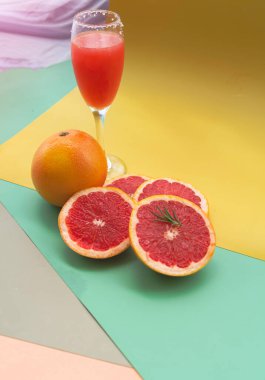 Renkli arka planda, meyve suyu bardağının önüne koyulmuş, orta pembe hamurlu greyfurt.