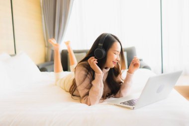 Güzel Asyalı bir kadının portresi. Yatak odasının içinde müzik dinlemek için kulaklıklı dizüstü bilgisayar kullanıyor.
