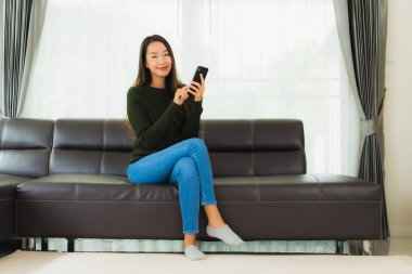 Güzel bir Asyalı kadın oturma odasındaki koltukta akıllı cep telefonu ya da cep telefonu kullanıyor.
