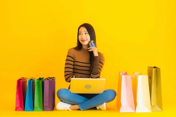 漂亮的肖像画年轻的亚洲女人带着笔记本电脑或电脑坐在地板上 用信用卡在黄色背景的购物袋里进行网上购物 — 图库照片