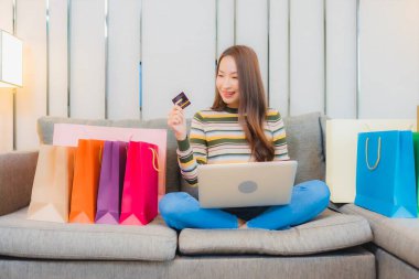 Asyalı güzel bir kadının portresi. Kanepede bilgisayar bilgisayarı, kredi kartı ve alışveriş çantası kullanıyor.