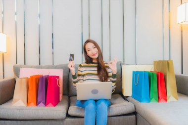 Asyalı güzel bir kadının portresi. Kanepede bilgisayar bilgisayarı, kredi kartı ve alışveriş çantası kullanıyor.