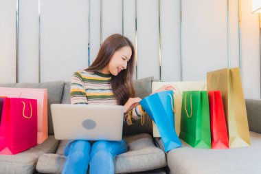 Asyalı güzel bir kadının portresi. Kanepede bilgisayar bilgisayarı, kredi kartı ve alışveriş çantası kullanıyor.