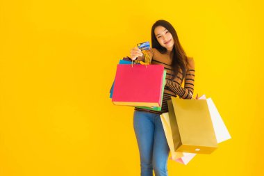 Güzel bir Asyalı kadının portresi sarı izole edilmiş arka planda renkli alışveriş torbalarıyla alışveriş yapmak için kredi kartı kullanıyor.