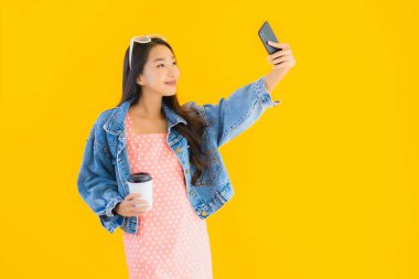 Kahve fincanı ve sarı izole edilmiş arka planda akıllı cep telefonlu güzel Asyalı kadın portresi.