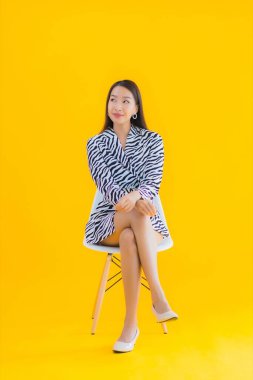 Güzel bir Asyalı kadın, sarı izole edilmiş arka planda hareketli bir şekilde sandalyede oturuyor.