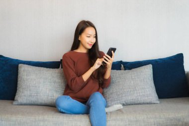 Portre güzel Asyalı kadın oturma odasındaki kanepede akıllı telefon kullanıyor.
