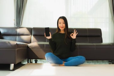 Güzel bir Asyalı kadın portresi oturma odasının iç tarafında kanepeli bir kahve fincanı ve akıllı cep telefonu kullanıyor.