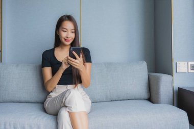 Güzel bir Asyalı kadın oturma odasındaki kanepede akıllı cep telefonu kullanıyor.