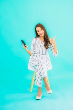 Güzel bir Asyalı kadın portresi akıllı cep telefonu kullanır ve mavi izole edilmiş arka planı olan bir sandalyeye oturur.