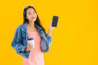 Kahve fincanı ve sarı izole edilmiş arka planda akıllı cep telefonlu güzel Asyalı kadın portresi.