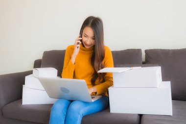 Asyalı genç bir kadının portresi, oturma odasındaki koltukta paket kutusuyla çalışmak için dizüstü bilgisayar kullanıyor.