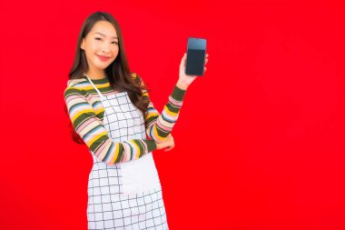 Güzel bir Asyalı kadın portresi kırmızı izole edilmiş arka planda akıllı cep telefonuyla önlük giyiyor.