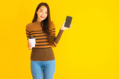 Güzel, genç Asyalı bir kadın sarı izole edilmiş arka planda kahve fincanı ile akıllı cep telefonu veya cep telefonu kullanır.