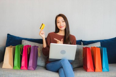 Asyalı güzel bir kadının portresi oturma odasındaki kanepede online alışveriş yapmak için bilgisayar ve kredi kartı kullanıyor.