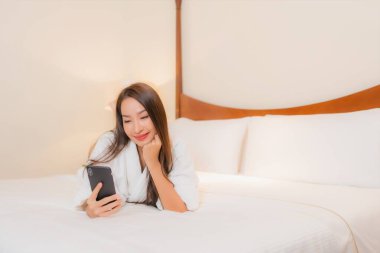 Güzel bir Asyalı kadın yatak odasının içinde akıllı cep telefonu kullanıyor.