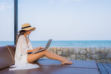 Plaj denizi çevresinde çalışmak için dizüstü bilgisayarı kullanan Asyalı genç bir kadının portresi.