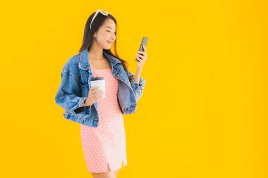 Kahve fincanı ve sarı izole edilmiş arka planda akıllı cep telefonlu güzel Asyalı kadın portresi.