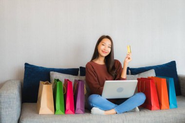 Asyalı güzel bir kadının portresi oturma odasındaki kanepede online alışveriş yapmak için bilgisayar ve kredi kartı kullanıyor.