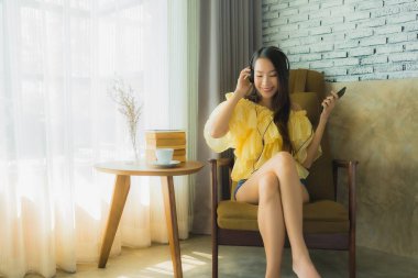 Genç Asyalı bir kadının portresi sandalyeye oturup oturma odasında cep telefonu kahvesi ve kitapla müzik dinlemek.