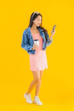 Kahve fincanı ve sarı izole edilmiş arka planda akıllı cep telefonlu güzel Asyalı kadın portresi.