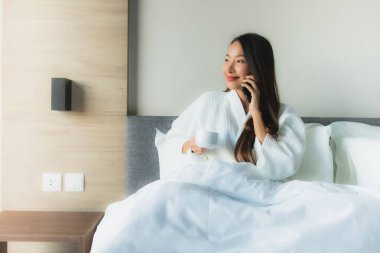 Güzel Asyalı genç kadınların portresi. Mutlu bir gülümseme. Yatak odasındaki yatakta kahve ve cep telefonuyla.
