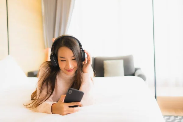 Portrait beautiful young asian woman using mobile phone with head phone for listen music in bedroom interior