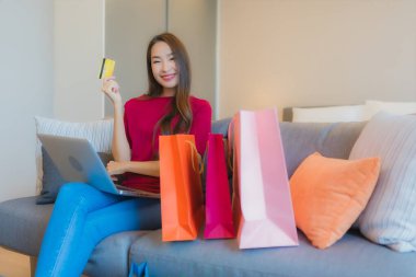 Güzel bir Asyalı kadın portresi. Kanepede çantalı online alışveriş için bilgisayarla kredi kartı kullanıyor.
