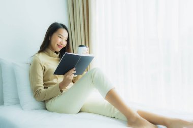 Genç Asyalı kadın, elinde kahve fincanıyla yatak odasında kitap okuyor.
