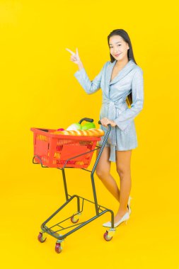 Asyalı güzel bir kadının portresi. Sarı arka planda süpermarketten alınmış alışveriş sepetiyle gülümsüyor.